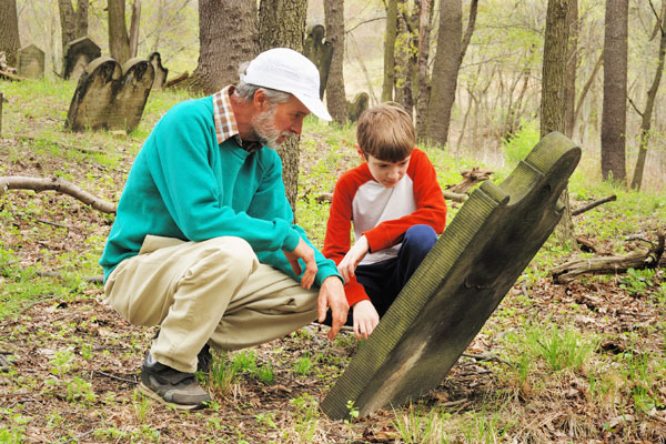 Older man and a younger boy in a wooded area with a tilted gravestone.